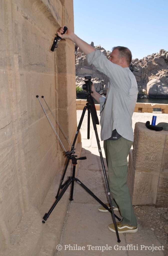 Image of Nick Hedley standing near one of the walls of the temple holding a light source in one hand while taking a photo of some graffiti with the help of a tripod. 
