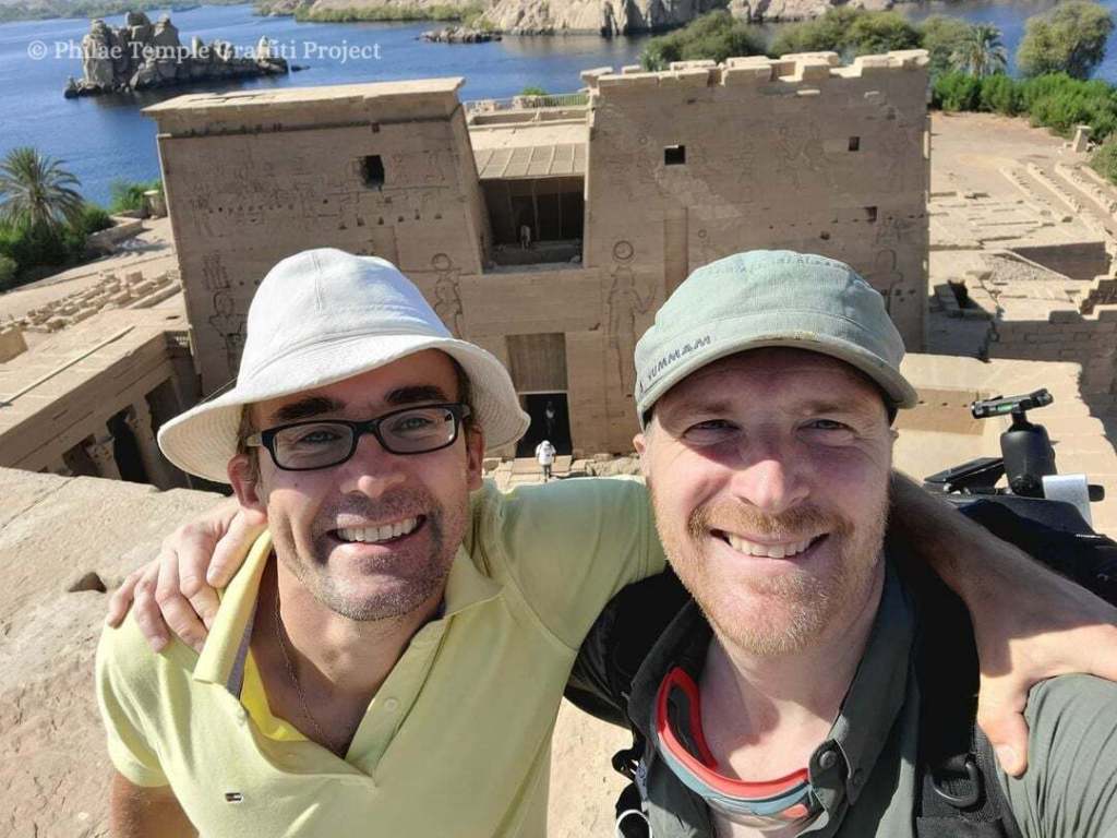 Photo of Dr. Jitse H.F. Dijkstra (left), wearing a yellow shirt and white bucket hat, and Dr. Nick Hedley (right), wearing a green shirt and green cap, standing on the roof of one of the Pylons at Philae. 